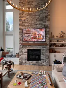 Samsung Frame TV mounted above a stone fireplace in a modern living room with tall windows and custom built in shelving.