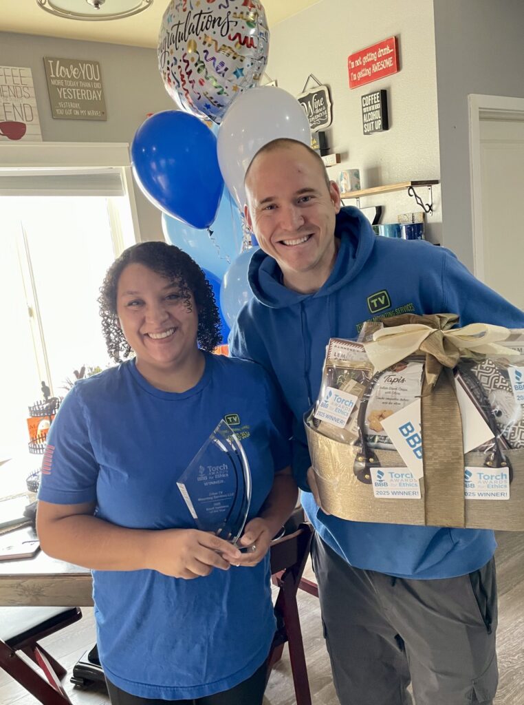 Alex and Jenae Zehm holding their BBB Torch Award trophy and celebration gift basket inside their home after winning the 2025 ethics award